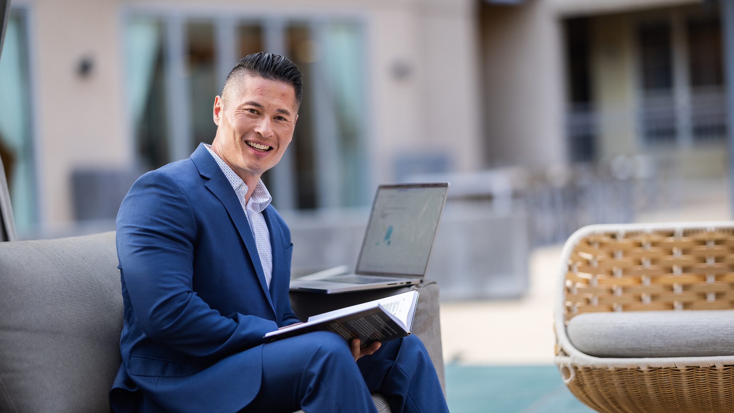 Man sitting outside holding book and working on laptop computer.